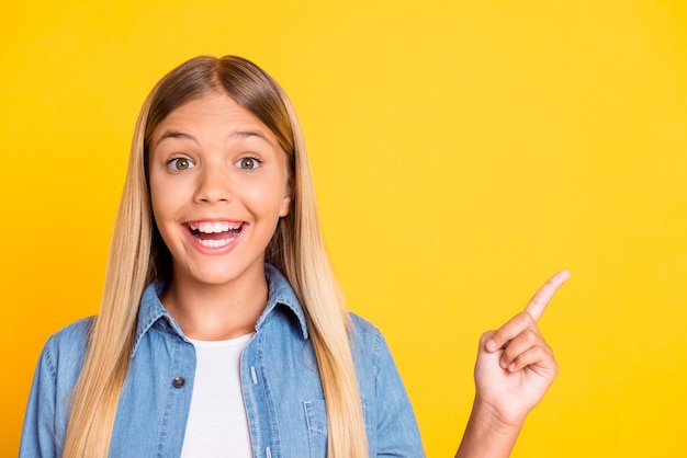 Foto de close up de uma loira engraçada pré-adolescente apontando para um espaço em branco usando uma camisa jeans isolada em um fundo de cor amarela brilhante