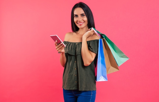 Foto de close-up de uma mulher moderna em uma roupa casual, sorrindo para a câmera, segurando um smartphone na mão direita e três sacolas de papel na mão esquerda