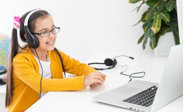 Foto de close-up de uma pequena aluna bonita senhora da escola falando skype notebook lição on-line videochamada sentar mesa usar fones de ouvido distância cumprimentar colegas acenar braço quarentena estudar sala de estar dentro de casa.