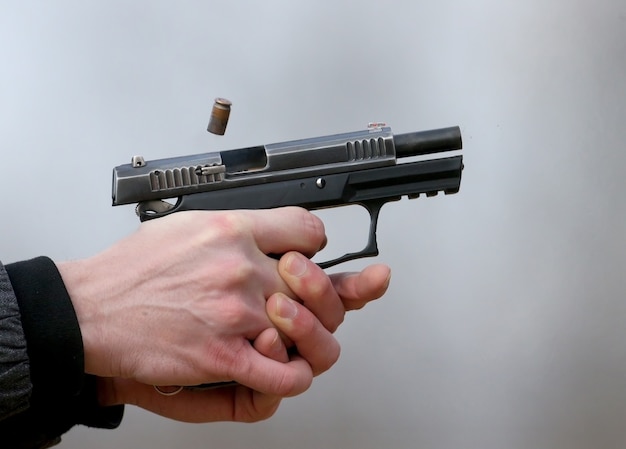 Foto de close-up de uma pistola disparando com as duas mãos, os projéteis emanando da veneziana e fumaça azul.