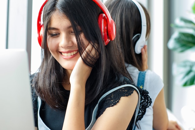 Foto de close-up retrato de um giro sorridente jovem adolescente tailandês-turco sentado de costas em um sofá vermelho e ouvindo música. Irmãs mestiças morando juntas e uma jogando laptop