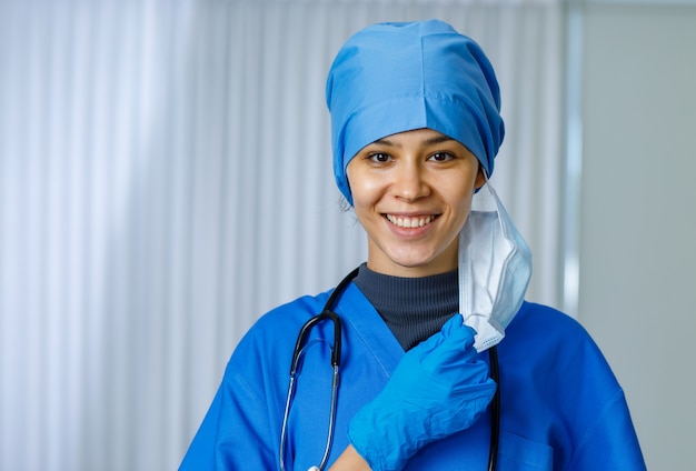 Foto de closeup retrato de feliz linda médica liberdade feminina usa terno hospitalar azul com estetoscópio sorrindo quando tira a máscara cirúrgica usada, olha para a câmera enquanto a pandemia de coronavírus termina