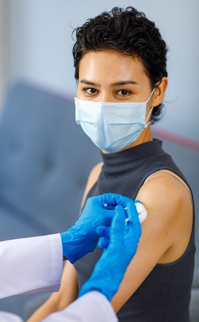 Foto de closeup retrato de paciente do sexo feminino usa máscara facial, sente-se olhando para a câmera enquanto o médico usa jaleco branco e luvas de borracha, usando agulha de seringa e vacina de injeção de coronavírus de algodão.