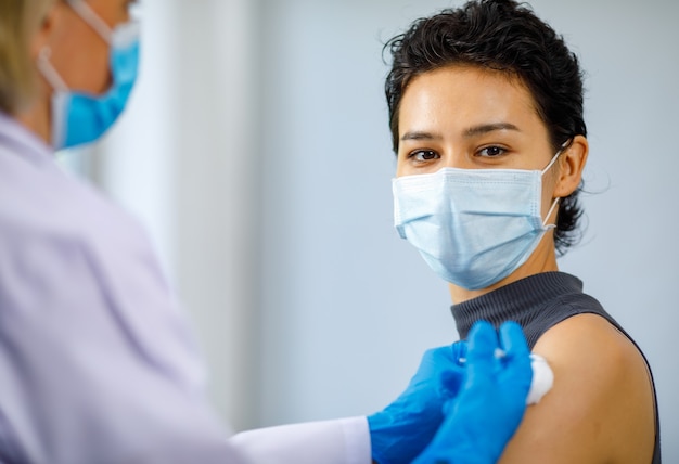 Foto de closeup retrato de paciente do sexo feminino usa máscara facial, sente-se olhando para a câmera enquanto o médico usa jaleco branco e luvas de borracha, usando agulha de seringa e vacina de injeção de coronavírus de algodão.