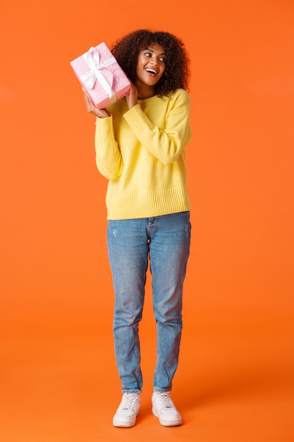 Foto de corpo inteiro curiosa adorável garota afro-americana receber presente de natal e interessar o que há dentro, sacudindo a caixa de presente adivinhe o que há, sorrindo comemorando as férias de inverno, ficou presente