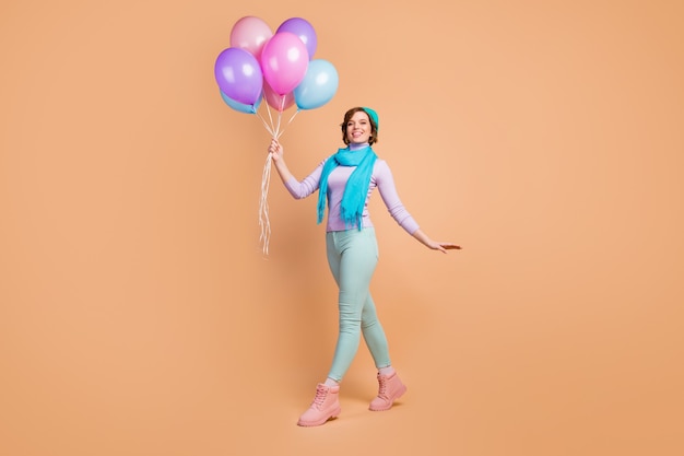 Foto de corpo inteiro da moça bonita carregando muitos balões de ar caminhando festa surpresa melhor amiga usar jumper violeta lilás calças verdes botas boina azul lenço isolado fundo de cor bege