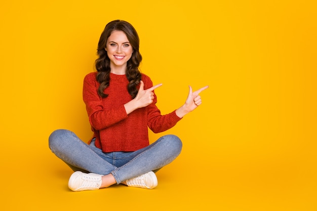 Foto de corpo inteiro da promotora garota confiante e positiva sentar-se no chão com as pernas cruzadas e apontar o dedo indicador para demonstrar a promoção de anúncios usar suéter jeans isolado sobre um fundo de cor brilhante