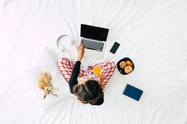 Foto de corpo inteiro da vista superior de uma jovem morena com roupas casuais, enquanto ela trabalha com um laptop na cama. Garota feliz estudando com um cachorro em casa