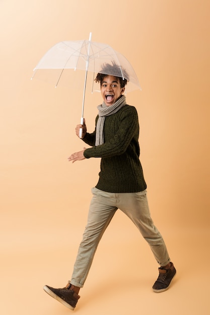 Foto de corpo inteiro de adolescente afro-americano vestindo blusa e cachecol andando sob o guarda-chuva, isolada sobre uma parede bege