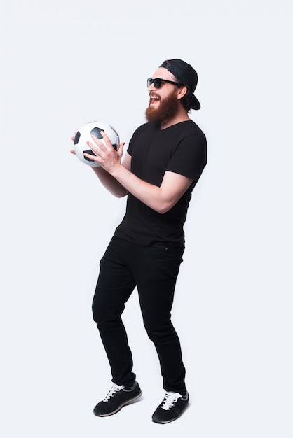 Foto de corpo inteiro de alegre jovem barbudo segurando bola de futebol e sorrindo sobre branco