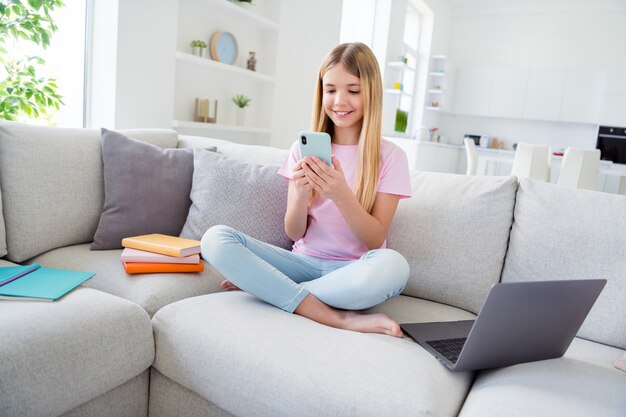 Foto de corpo inteiro de criança positiva menina estudo remoto sentar conforto sofá pernas cruzadas usar smartphone comentário rede social postar em casa dentro de casa