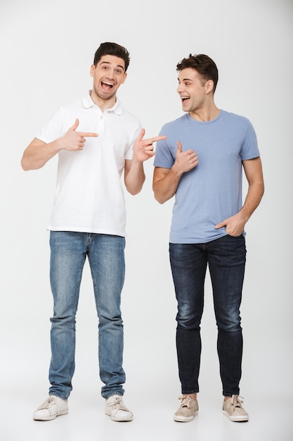 Foto de corpo inteiro de dois homens bonitos dos anos 30 vestindo camiseta casual e jeans, sorrindo e gesticulando os dedos uns contra os outros, isolado sobre fundo branco