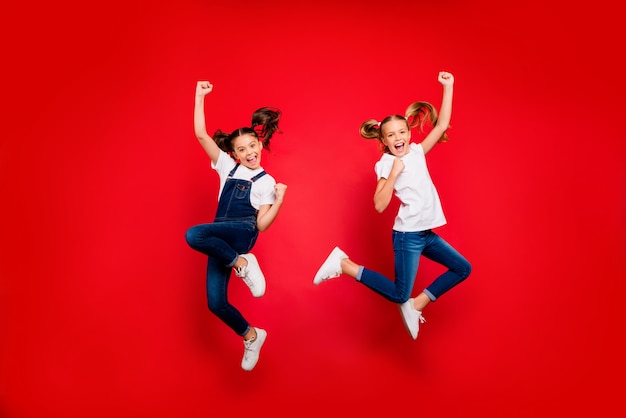 Foto de corpo inteiro de duas namoradas pequenas e alegres celebram o triunfo levantam os punhos gritam sim, pule vestir camiseta branca jeans jeans isolado tênis de fundo colorido