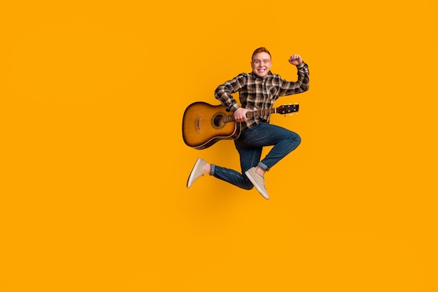 Foto de corpo inteiro de jovem saltador ativo celebrando punhos de sorte tocando concerto de guitarra isolado sobre fundo de cor amarela