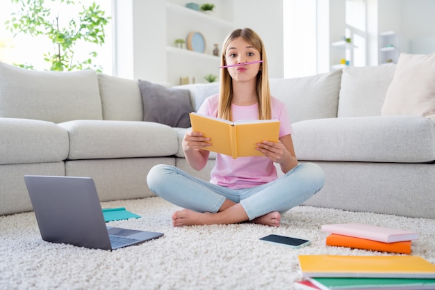 Foto de corpo inteiro de menina louca criança estudo remoto escrever redação caderno preciso descansar relaxar colocar nariz lápis careta rosto sentar chão tapete pernas cruzadas dobradas em casa dentro de casa