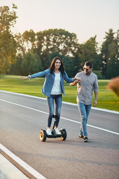 Foto de corpo inteiro de mulher feliz andando de hoverboard elétrico na estrada com namorado segurando a mão dela