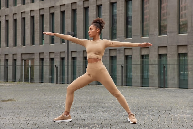 Foto de corpo inteiro de mulher magra e esportiva mantendo os braços de lado, exercícios ao ar livre vestida com leggings cortadas e poses de tênis ao ar livre