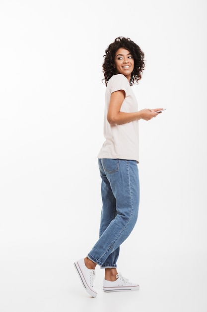 Foto foto de corpo inteiro de mulher morena atraente com penteado afro, vestindo camiseta e jeans usando smartphone e olhando para trás, isolado sobre a parede branca