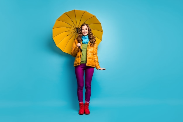 Foto de corpo inteiro de senhora bonita engraçada segurando guarda-chuva brilhante aproveite dia ensolarado de primavera caminhada pela rua no exterior usar casaco amarelo lenço calça violeta sapatos vermelhos parede cor azul