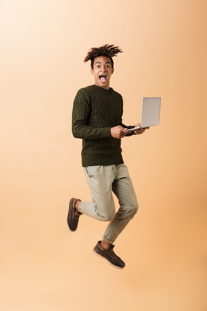 Foto de corpo inteiro de um afro-americano satisfeito com um suéter andando com o laptop, isolada sobre uma parede bege