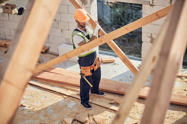 Foto de corpo inteiro de um construtor totalmente equipado em um capacete de segurança carregando madeira para construção