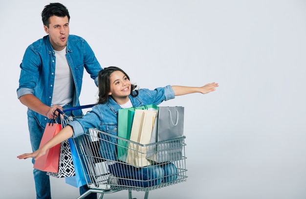 Foto de corpo inteiro de um homem atraente e feliz em uma roupa casual, andando em um carrinho de compras com sua filha sorridente, que está imitando um avião, e três sacolas de compras dentro.