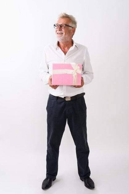 Foto de corpo inteiro de um homem barbudo sênior feliz, sorrindo e pensando, segurando uma caixa de presente em branco