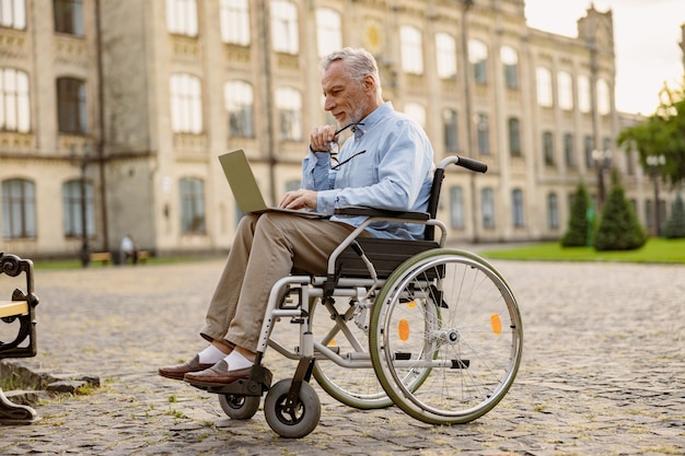 Foto de corpo inteiro de um homem maduro recuperando um paciente em cadeira de rodas trabalhando em um laptop ao ar livre