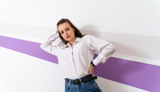 Foto de corpo inteiro de uma garota em uma camisa branca e calça jeans. mulher está de pé perto de uma parede branca com faixa violeta. mão sobre a cabeça.