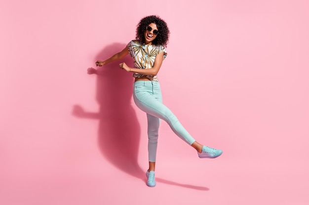 Foto de corpo inteiro de uma garota engraçada feliz de pele negra dançando no chão, chutando, isolada em um fundo de cor rosa pastel