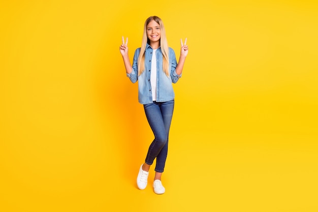 Foto de corpo inteiro de uma garota loira pré-adolescente sorrindo alegremente, mostrando o sinal V com as duas mãos usando roupas jeans isoladas em um fundo de cor amarelo brilhante