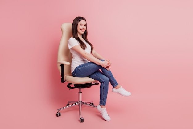 Foto de corpo inteiro de uma jovem feliz e sorridente, de camiseta e jeans, sentada em uma cadeira, isolada sobre um fundo de cor rosa