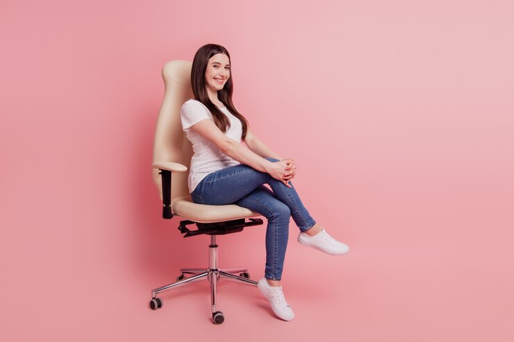 Foto foto de corpo inteiro de uma jovem feliz e sorridente, de camiseta e jeans, sentada em uma cadeira, isolada sobre um fundo de cor rosa