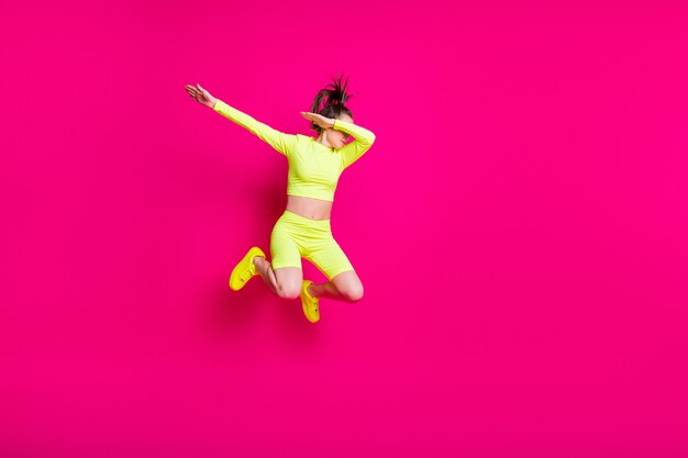 Foto de corpo inteiro de uma jovem saltitante vestindo roupas esportivas amarelas, mostrando o hype isolado em um fundo de cor rosa brilhante