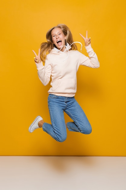 Foto de corpo inteiro de uma linda garota da escola com aparelho dentário pulando e mostrando o símbolo da paz, isolada sobre fundo amarelo