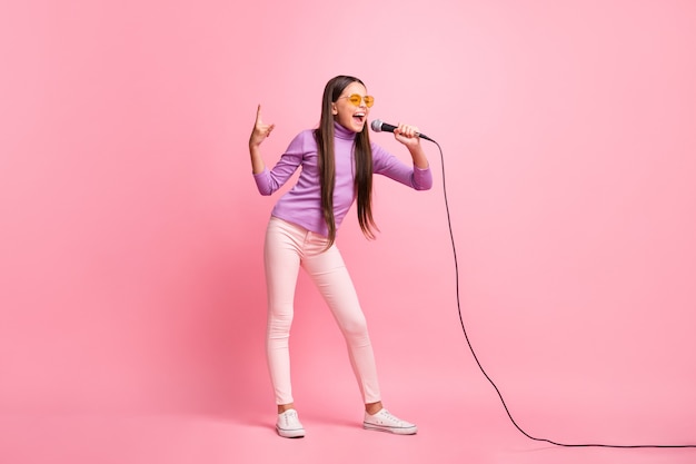 Foto de corpo inteiro de uma menina pequena cantando música no microfone usando calças suéter violeta isoladas sobre um fundo de cor pastel