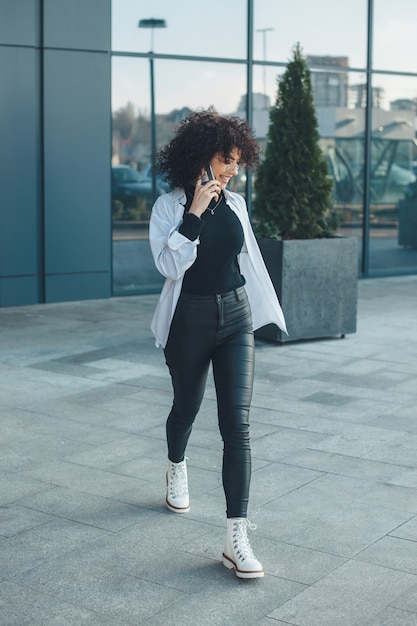 Foto de corpo inteiro de uma mulher caucasiana com cabelo encaracolado falando ao telefone do lado de fora e sorrindo