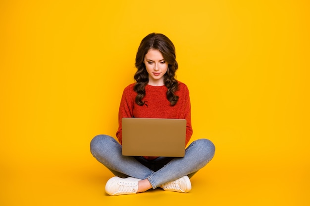 Foto de corpo inteiro de uma senhora atraente encaracolada de bom humor sentado no chão trabalhando com o computador ficar em casa. Trabalhador remoto usar jeans azul de malha vermelha suéter isolado fundo de cor amarela brilhante