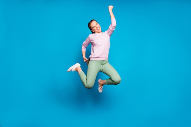 Foto de corpo inteiro de uma senhora muito louca de tranças longas pulando campeã de competições esportivas usando calças casuais rosa pulôver verde com fundo de cor azul isolado