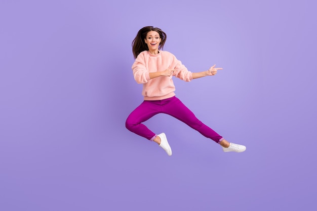 Foto de corpo inteiro de uma senhora pulando alto, indicando um espaço vazio com os dedos na parede roxa