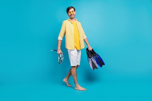 Foto de corpo inteiro positivo cara turista vá caminhar praia tente esportes aquáticos segurar máscara óculos nadadeiras tubo usar camisa amarela listrada branca curta descalça pé isolado azul cor de fundo