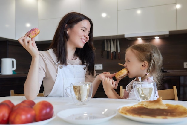 Foto de corte da mãe de vermelho e sua filha com donuts coloridos sentados na cozinha Conceito de dieta e junk food