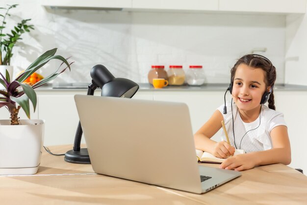 Foto de criança positiva menina estudo remoto sentar mesa mesa usando laptop tem aula de comunicação online com tutor redigindo relatório em casa dentro de casa