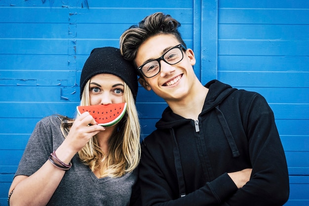 Foto de dois amigos em pé pagando juntos - ela está brincando com melancia - ele está de óculos