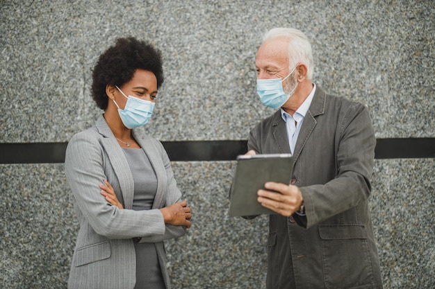 Foto de dois empresários multiétnicos bem-sucedidos com máscara protetora usando tablet digital e discutindo em pé contra uma parede de prédio corporativo durante a pandemia de COVID-19.