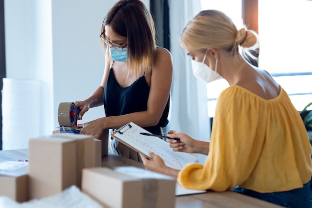 Foto de duas belas vendedoras freelance de mulheres de negócios usando uma máscara facial higiênica enquanto verificava o pedido do produto enquanto embalava e lacrava caixas de papelão em sua pequena empresa inicial.