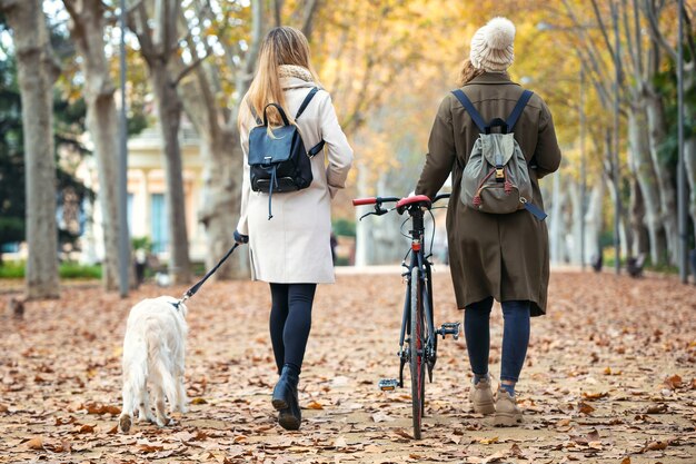 Foto de duas lindas amigas caminhando e andando de bicicleta no parque enquanto vão com o cachorro no parque no outono.