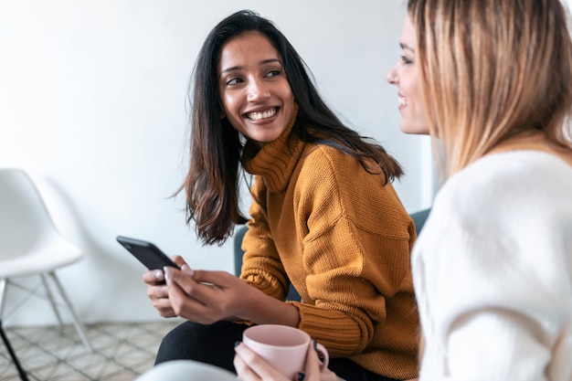 Foto de duas mulheres bonitas usando o celular enquanto bebia café no sofá em casa.