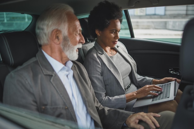 Foto de duas pessoas multiétnicas bem-sucedidas usando laptop e conversando sobre trabalho enquanto estão sentadas no banco de trás de um carro durante o trajeto de negócios.