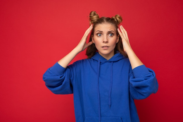 Foto de emocional surpresa espantada jovem mulher loira bonita com dois chifres com emoções sinceras, vestindo moderno capuz azul brilhante isolado sobre um fundo vermelho com espaço livre.